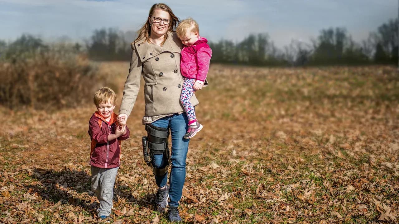 A mother holds one child while holding hands with another as they stroll outside through a field. The mother is wearing Ottobock's C-Brace.